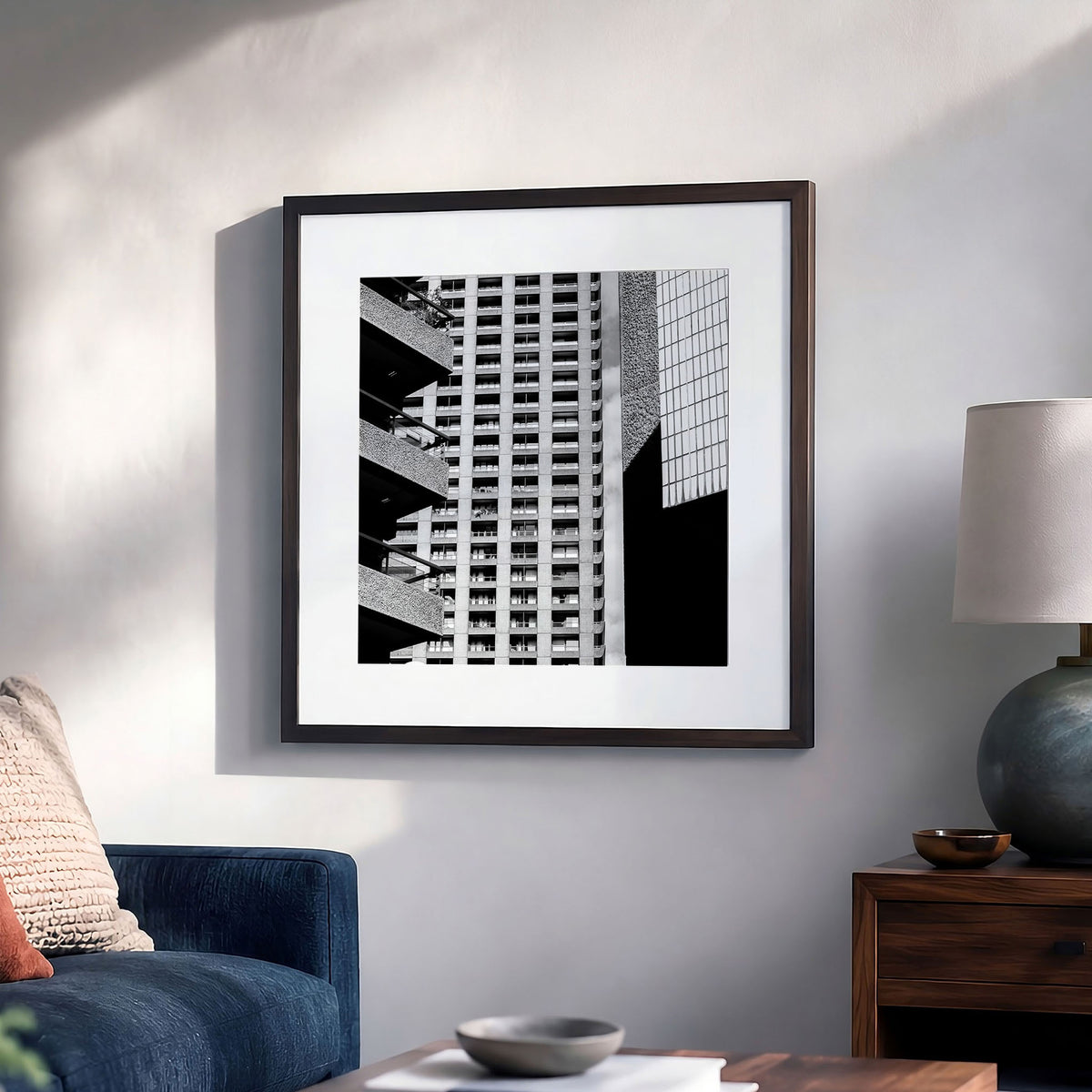 Black and white architectural art print of a modern building facade, displayed in a light brown frame on a light gray wall in a styled living room setting with a blue sofa, wooden side table, and lamp.
