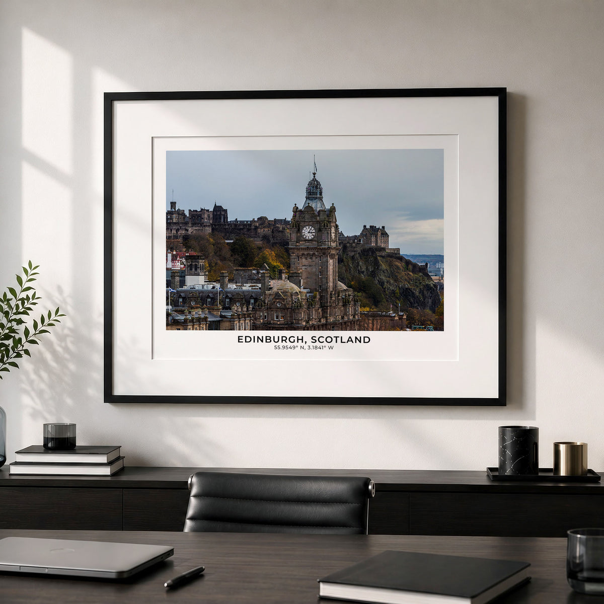 A framed art print of Edinburgh, Scotland, featuring the historic Edinburgh Castle and Old Town, styled on a light gray wall above a dark wooden desk with a laptop and other office items.
