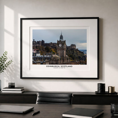 A framed art print of Edinburgh, Scotland, featuring the historic Edinburgh Castle and Old Town, styled on a light gray wall above a dark wooden desk with a laptop and other office items.