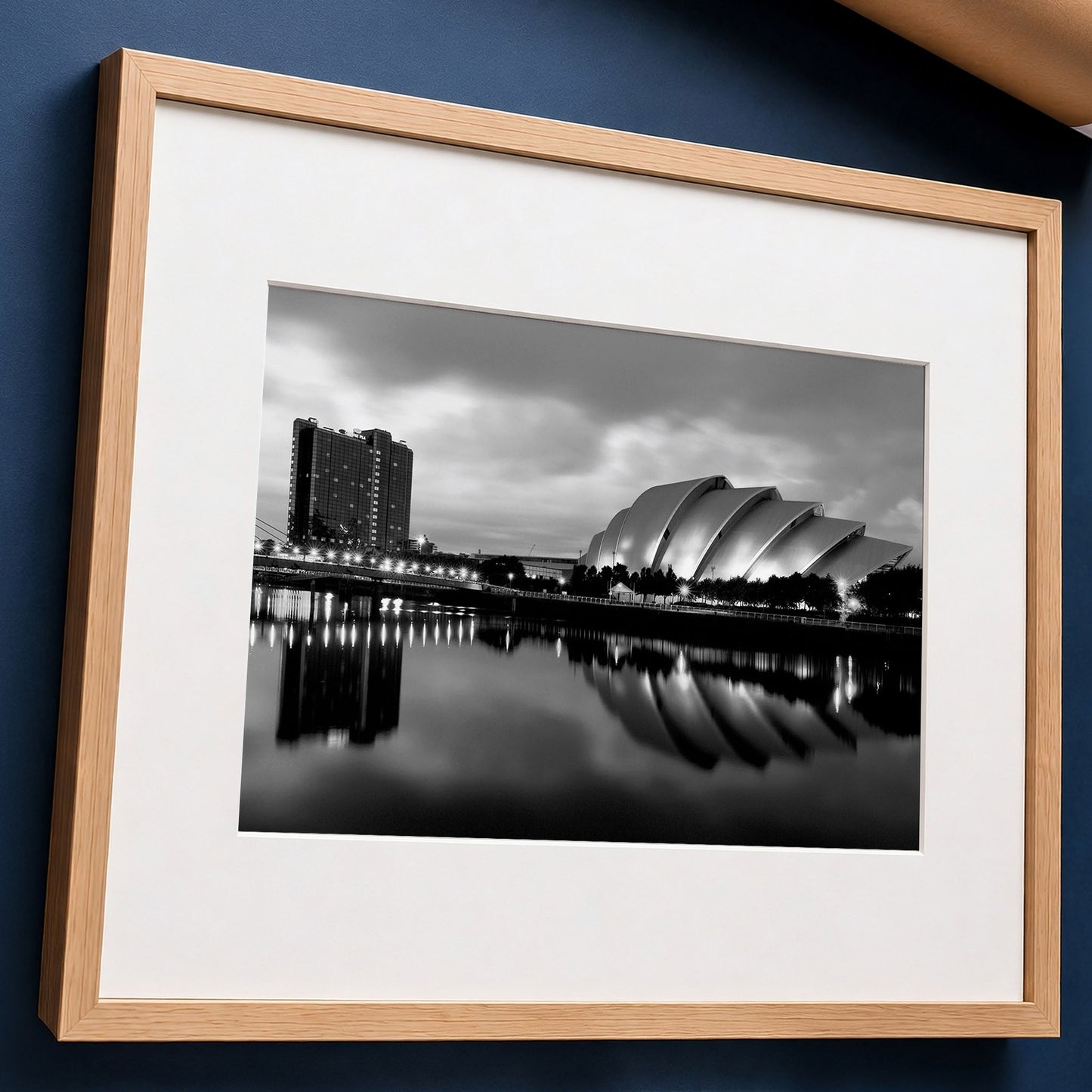 A framed black and white photograph of the Armadillo in Glasgow reflected in the River Clyde, presented in a light wood frame with a white photo mount, laid flat against dark blue surface.
